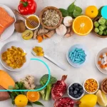 این تصویر ترکیبی از مکمل‌های غذایی (کپسول‌ها و قرص‌ها) و غذاهای عملکردی طبیعی مانند ماهی سالمون، زردچوبه، عسل، کیوی، مرکبات، مغزها و سبزیجات را نشان می‌دهد که برای تغذیه مناسب بعد از ورززش برای ریکاوری سریع است